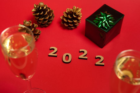 A Christmas decoration with wooden numerals, 2022 year, Christmas gift in wrapping paper with shiny glitter green bow, golden pine cones and champagne flutes with a sparkling wine on a red backgroundの写真素材