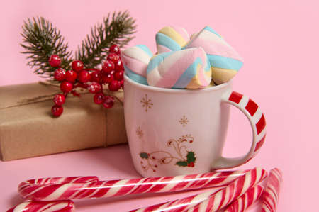 Close-up of coffee mug with colorful marshmallows, Christmas gift in craft wrapping paper, holly and...