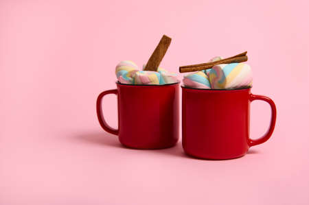 Marshmallows and cinnamon stick in cup of hot chocolate drink in red mugs isolated over pink pastel...