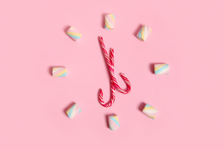 Top view with colorful sweet marshmallows and striped Christmas lollipops, candy canes stacked in the shape of a clock and dial, isolated on pink pastel colored background with copy space for adの写真素材