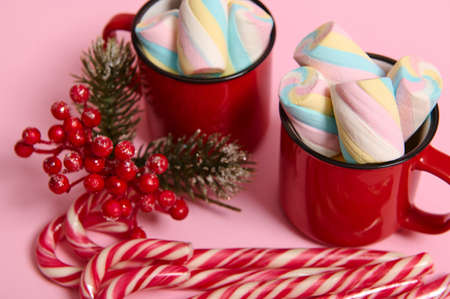 Close-up, cropped image of Christmas items, striped sugary candy canes, holly and mug with hot drink and wet colorful marshmallows on pink colored background with copy space for Christmas adの写真素材