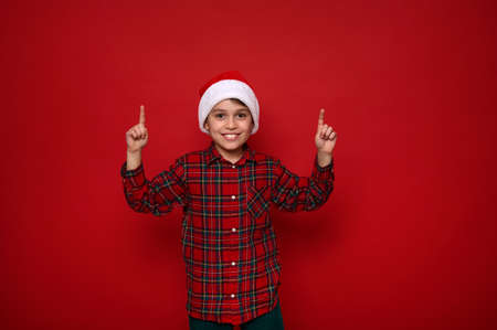 Adorable European preadolescent cheerful boy n Santa hat and checkered shirt points with fingers up on a copy space on red colored background for Christmas and New Year advertisementの写真素材