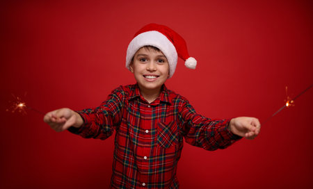 Handsome adorable preteen boy, pretty child in Santa Claus hat and checkered shirt enjoys the Christmas party, plays with Bengal lights, sparklers, isolated over red background with copy space for adの写真素材