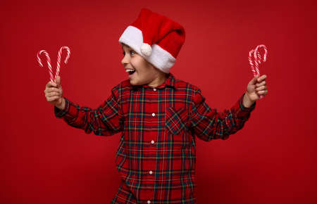 Beautiful child boy in checkered shirt and Santa Claus hat looking at Christmas lollipops, striped...