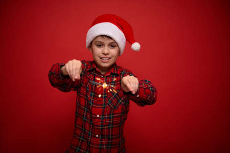 Handsome adorable preteen boy, beautiful child in Santa Claus hat and checkered shirt enjoys the Christmas party, plays with Bengal lights, sparklers, isolated o red background with copy space for adの写真素材
