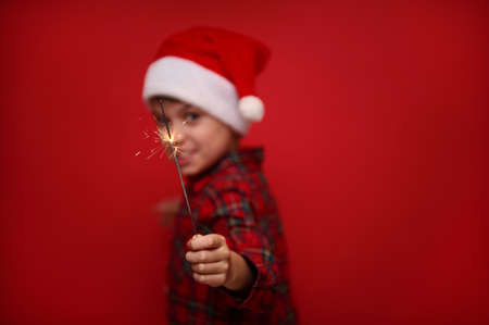Focus on the fire sparkles in the sparklers bengal lights in the hand of blurred cute boy in Santa Claus hat and checkered shirt, isolated over red colored background with copy space for Christmas adの写真素材