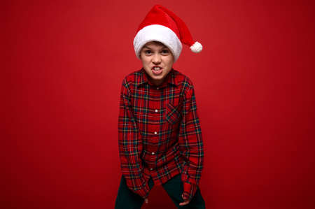Funny preadolescent boy in plaid shirt and Santa Claus hat grimacing looking at camera posing against red colored background with copy space for New Year and Christmas adの写真素材