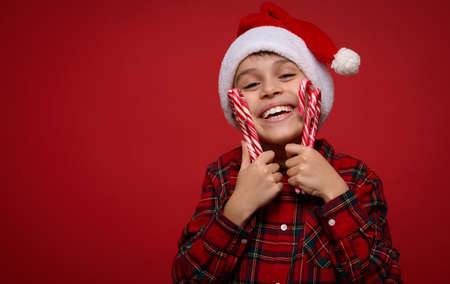 Headshot of adorable tender cute little boy, adorable smiling child in Santa hat and checkered shirt gently hugs Christmas lollipops sweet striped candy canes, isolated on red background, copy spaceの写真素材