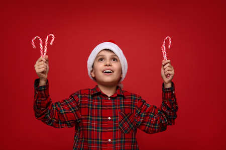 Adorable 9 years old child boy in Santa hat holds sweet striped Christmas lollipops, candy canes in hands and smiles cutely with a beautiful toothy smile, looking into copy space on a red background.の写真素材
