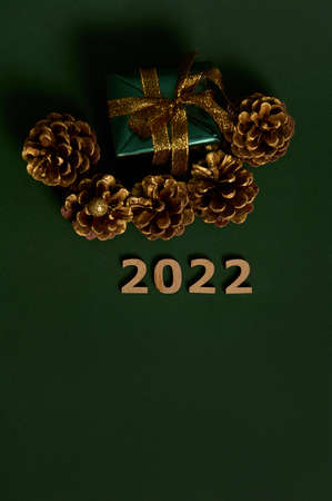 Top view of Christmas present in glitter wrapping paper with golden bow and pines cones colored in gold, wooden numerals symbolizing the year 2022 laid out on dark green background with copy spaceの写真素材