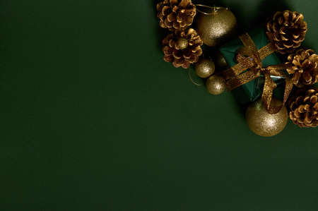 High angle view of a present in shiny green glitter wrapping gift paper with golden bow, pines cones and gold Christmas tree toys laid out on dark background. Copy space for advertisementの写真素材