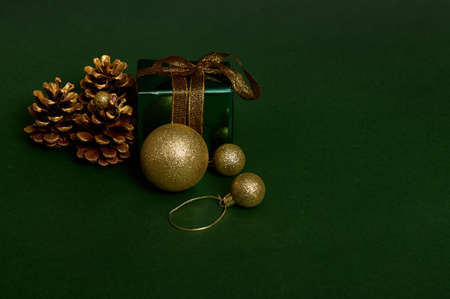 Front view of a present in shiny green glitter wrapping gift paper with gold ribbon and golden bow, pines cones and spherical Christmas tree toys laid out on dark background. Copy space for adの写真素材