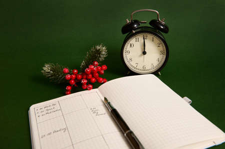 Close-up of an opened page of notebook, agenda with plans for day, alarm clock and snowy branch of holly Christmas plant isolated on green backgroundの写真素材