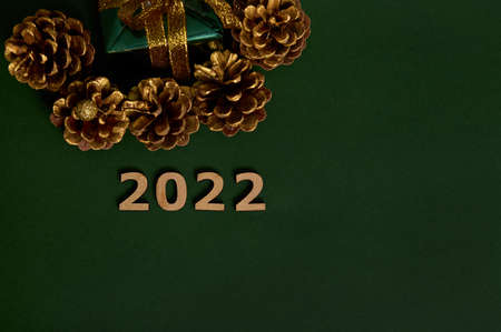 Top view of cropped Christmas present in green wrapping paper with golden bow and pines cones colored in gold, wooden numerals symbolizing the year 2022 laid out on dark background. Copy space for adの写真素材