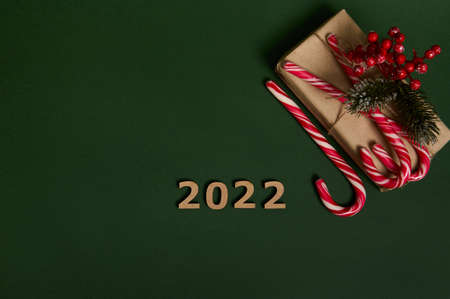 Flat lay of Christmas present in craft wrapping paper ornate with holly and sugary striped lollipop candy canes, wooden numerals 2022 laid out in the corner of dark green background with space for adの写真素材