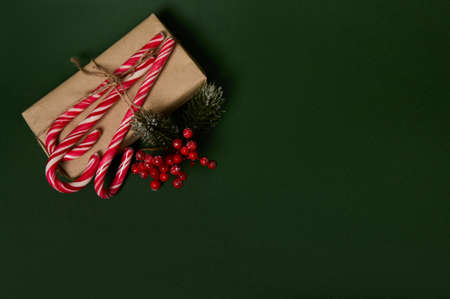 Copy space for advertisement on a dark green background with Christmas present in craft wrapping gift paper decorated with holly and sweet sugary striped white and red lollipops candy canesの写真素材