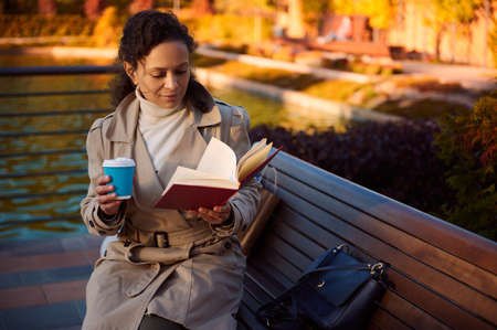 Young pretty woman in cozy warm outfit reads book and drinks coffee sitting on a wooden bench in...