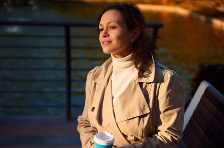 Portrait of young beautiful African woman in beige cozy outfit sitting on a park bench with cup of...