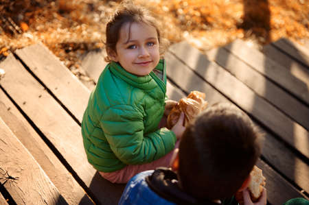 Overhead view of adorable girl wearing green jacket looking at camera sitting next to her brother with a baked delicious croissant in hand and enjoys a breakfast outdoor at autumnal nature backgroundの写真素材