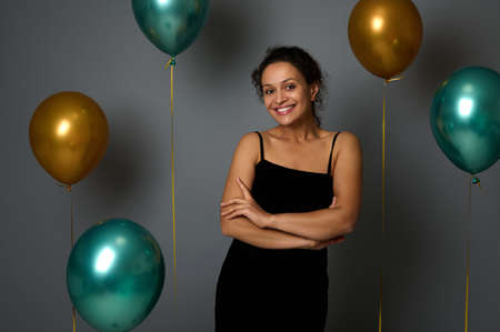 Gorgeous elegant Hispanic woman in a black evening dress posing with crossed arms, smiling cutely, looking at the camera on a gray background decorated with festive gold and green metal air balloonsの写真素材