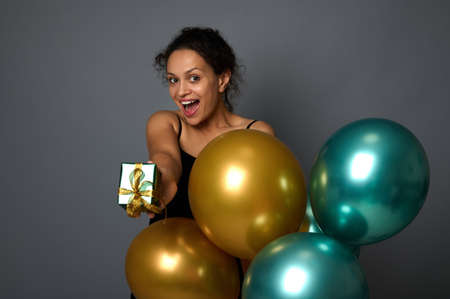 Smiling African woman showing a luxury Christmas gift box with golden bow and shiny metallic gold...