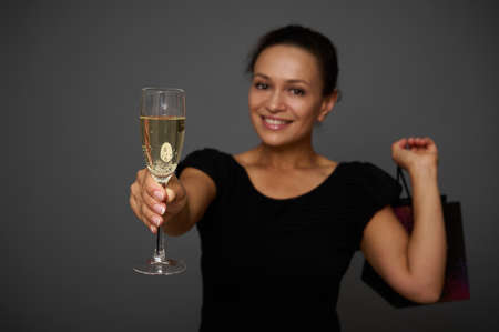 Soft focus on champagne flute, glass with sparkling wine in the hands of blurred smiling beautiful woman dressed in black and holding a black shopping bag, celebrating Black Friday. Copy space for adの写真素材