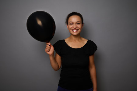 Cheerful African woman dressed in black smiles looking at camera and poses against gray background with a black colored air balloon in hand. Black Friday concept with copy space for adの写真素材