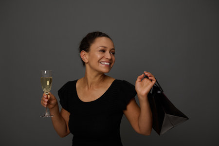 Beautiful woman shopper dressed in black dress smiles looking at copy space on a gray background with a glass of sarkling wine and shopping black packet. Concept of celebration the Black Fridayの写真素材