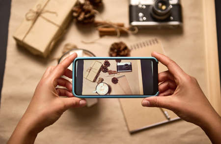 Human hands holding smartphone in live view mode and taking a photo of Christmas composition of cup with cocoa and marshmallows, vintage camera, craft gift box and pine cones on cardboard surfaceの写真素材