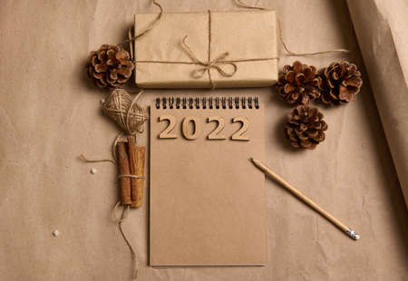 Flat lay arranged composition of a Christmas gift in craft wrapping paper, pine cones, cinnamons, rope and a pencil on notepad with empty blank brown paper on a cardboard background with copy ad spaceの写真素材