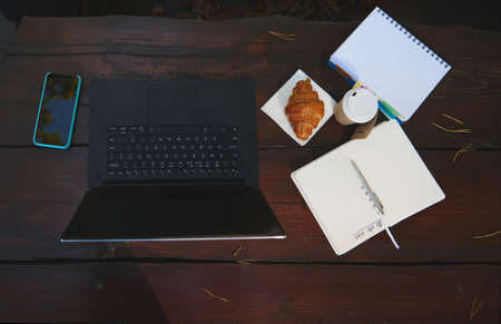 High angle view composition of business and office items. Laptop, smartphone, open copybook, notepad and fresh baked croissant with takeaway paper mug of hot coffee on a wooden table surface. Flat layの写真素材