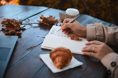 Close-up of the hand of a cropped woman holding silver pen and writing the list to do in notepad, while sitting at a wooden table with fallen dry oak leaves, takeaway paper cup and baked croissantの写真素材