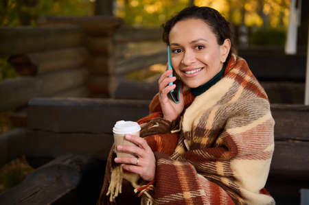 Pretty woman looks at camera talking on mobile phone, holding cardboard cup of hot drink, keeping warm while sitting on wooden bench wrapped in a cozy checkered woolen blanket on a cool autumn dayの写真素材