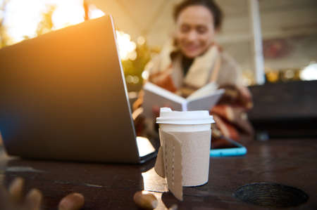 Takeaway cardboard paper cup with hot drink on table with laptop and blurred happy woman enjoying distant learning work outdoors at beautiful autumn sunset. Autumn background with copy spaceの写真素材