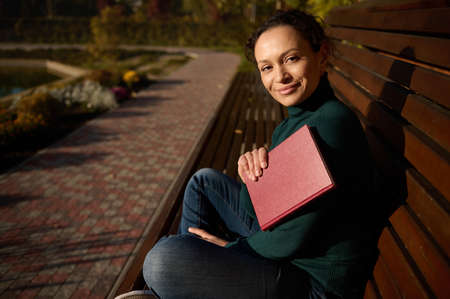 Gorgeous self-confident woman in casual denim rests in a park sitting in lotus position on a wooden...