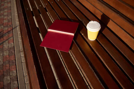 Top view of an open book with dark red hard cover and a yellow takeaway cardboard paper cup with hot drink on a wooden bench in the park. Concept of outdoor rest away from gadgets and city bustle.の写真素材