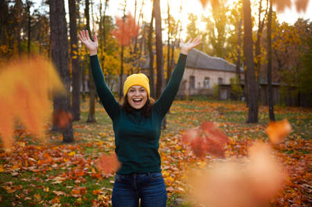 Lifestyle portrait of attractive lovely optimistic cheerful woman in warm woolen cozy clothes plays throwing colorful oak and maple dry autumn fallen leaves in the air enjoying walk in a city parkの写真素材