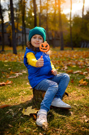 Happy Halloween concept. Adorable little child girl sitting on a tree stump in beautiful park at autumn sunset with falling sunbeams through trees, showing a baked gingerbread pumpkin to the camera.の写真素材
