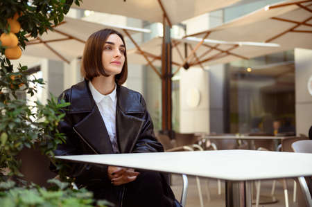 Beautiful pensive brunette sitting at table on summer terrace of a cozy cafe near tangerine decorative tree, looking thoughtfully aside waiting for an order and a meeting with business partnerの写真素材