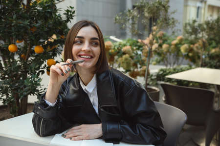 Lifestyle portrait of attractive fashionable brunette pretty woman in eco leather jacket holding pen...