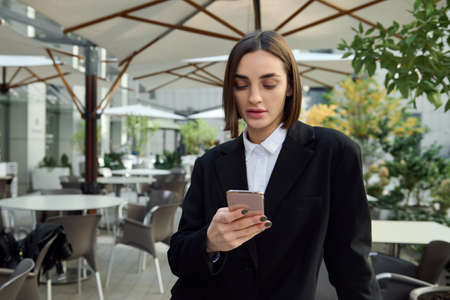 Self-confident woman holding mobile phone smartphone, typing text messages, messaging, swiping web page. Business, finance, online remote work, online shopping, communication and connection conceptsの写真素材
