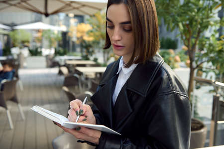 Close-up confident portrait of concentrated young business woman, copywriter, journalist, writing on white notepad while sitting outdoors on urban public background.の写真素材