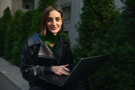 Close-up portrait of a young cute woman freelancer, copywriter, motivated start-up sitting on parapet with laptop, working remotely and smiles looking at camera. Connection and online business conceptの写真素材
