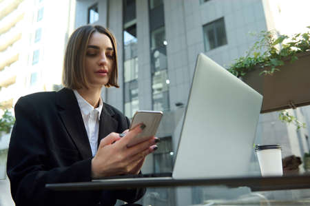 Self-confident motivated young freelance, business woman using mobile phone and working on laptop sitting in outdoor on urban background. Effectiveness, online business conceptの写真素材