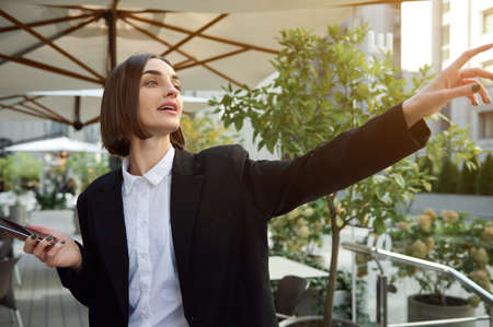 Cheerful young brunette woman holding smartphone, smiling toothy smile greeting while resting in open air on beautiful warm sunny day with sunrays falling on the backgroundの写真素材