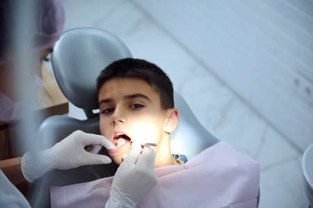 Overhead view of pediatric dentist examining oral cavity of a teenage boy sitting on dental chair, examines the teeth of the presence of caries using a dental mirror. School boy at dental appointmentの写真素材