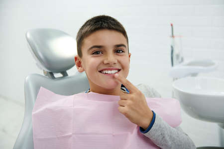 Happy boy holds finger near his mouth, looks at camera, smiles with beautiful toothy smile after...