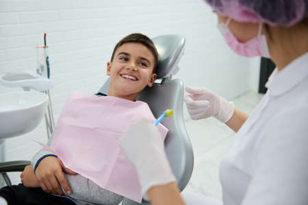 Handsome boy sitting in dentist chair smiles with beautiful toothy smile looking at a pediatric dentist teaching him about oral hygiene during dental appointment in white clean modern medical cabinetの写真素材