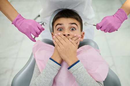 Frightened boy covers his mouth with his hands, sitting on dentist's chair on the background of hands in pink medical gloves, holding near his face a stainless steel mirror and forceps to remove teethの写真素材