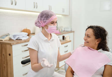 Dentist hygienist, dental assistant wearing pink medical mask and surgical gloves preparing woman patient to medical check-up, giving consultation about oral hygiene in dentistry contemporary clinicの写真素材